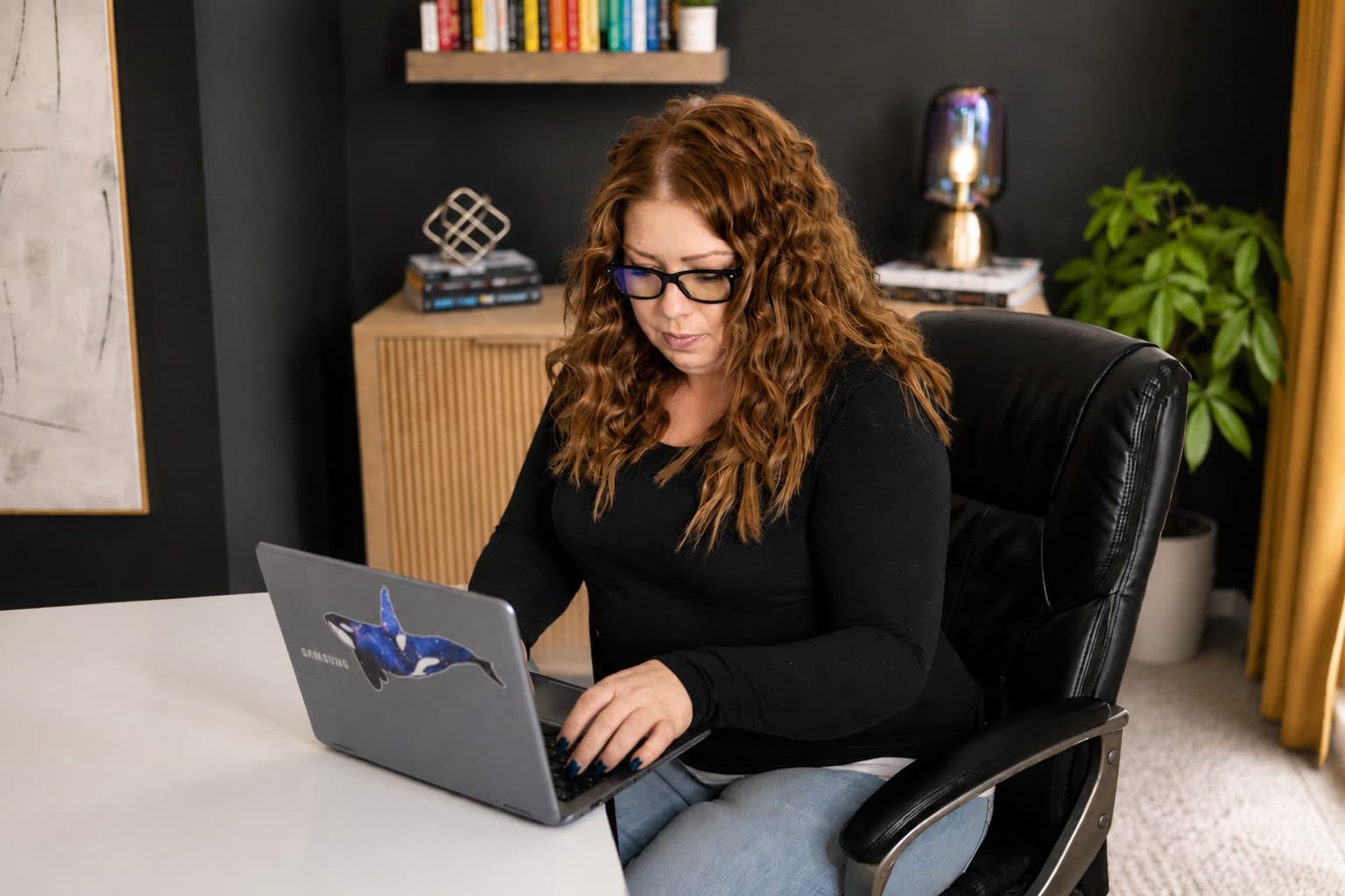 Racheal Blackmore writing at her laptop in her studio, lamp lit behind her.