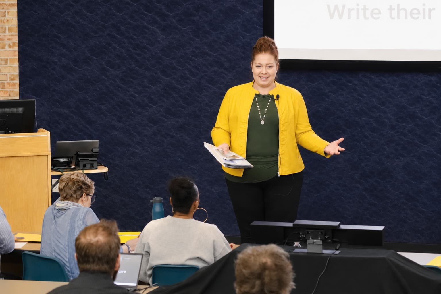Racheal Blackmore speaking to a workshop audience, gesturing mid-talk in a yellow jacket.